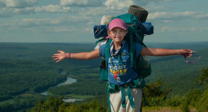 A person wearing a backpack stands high above a vast green forest below and spreads their arms in celebration 